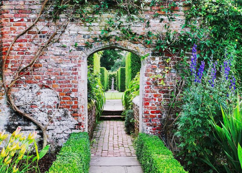 Archway in a brick wall with flowers and a statue.