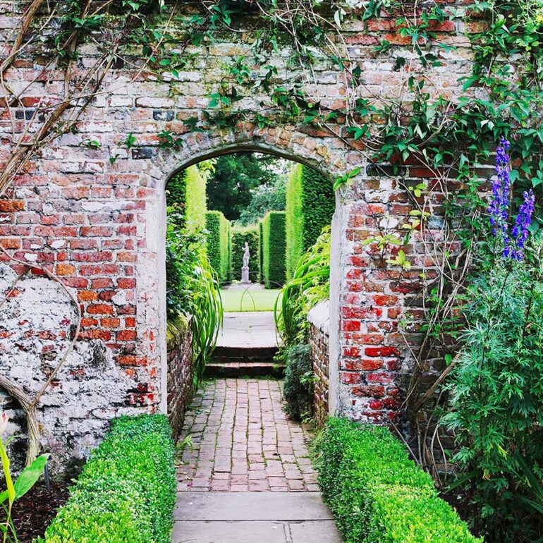 Archway in a brick wall with flowers and a statue.