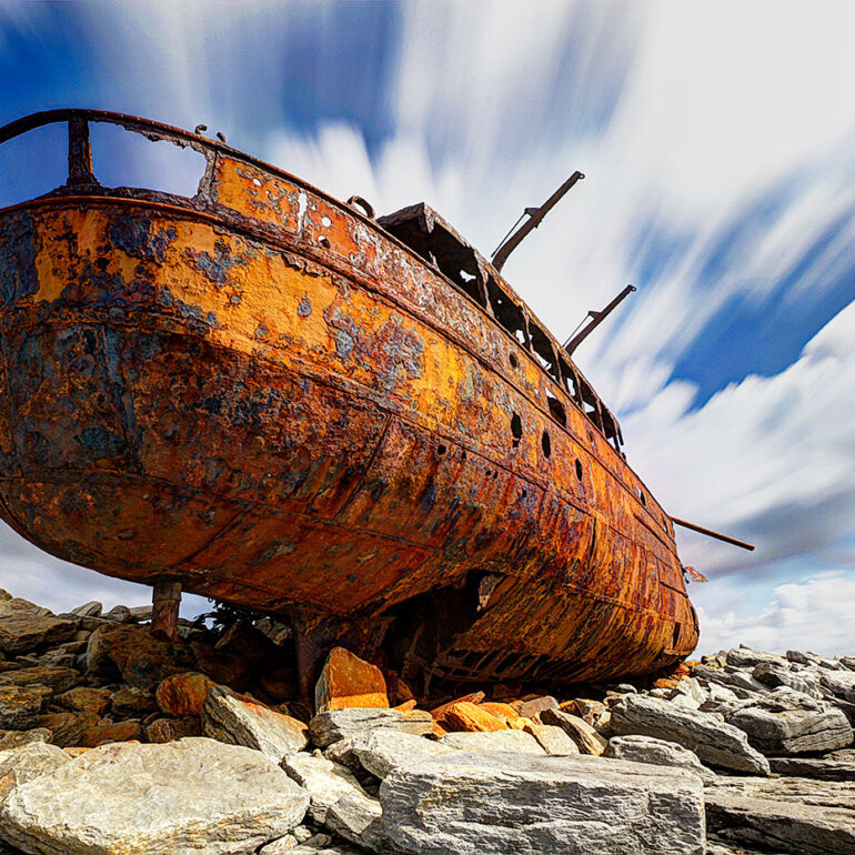 Rusted ship wreck on shore with vibrant clouds.