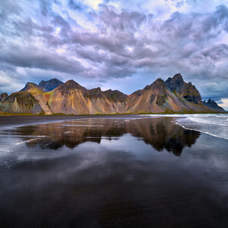 Mountain peaks reflected in the water along a black beach.